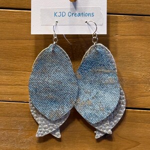 May include: A pair of layered earrings with a denim-like blue and silver design. The earrings are made of a textured material and have a fish-tail shape. The earrings are hanging from a white tag that reads "KJD Creations".