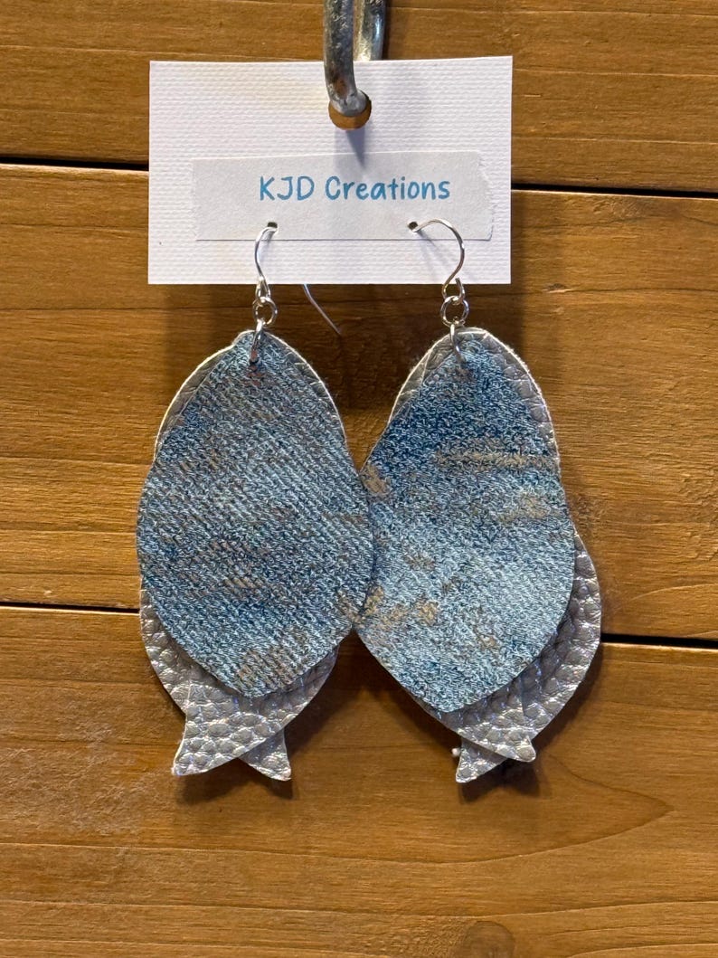 May include: A pair of layered earrings with a denim-like blue and silver design. The earrings are made of a textured material and have a fish-tail shape. The earrings are hanging from a white tag that reads "KJD Creations".
