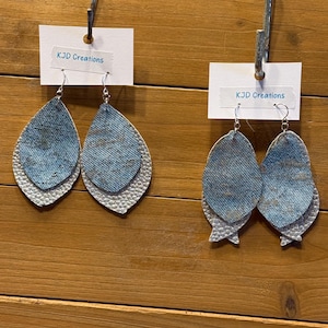 May include: Two pairs of layered teardrop earrings. The top layer is denim-like blue, and the bottom layer is white with a textured, scale-like pattern. Each earring hangs from a silver-colored hook. The text "KJD Creations" is on a white tag.