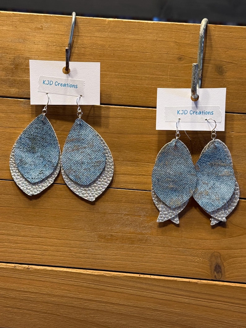 May include: Two pairs of layered teardrop earrings. The top layer is denim-like blue, and the bottom layer is white with a textured, scale-like pattern. Each earring hangs from a silver-colored hook. The text "KJD Creations" is on a white tag.