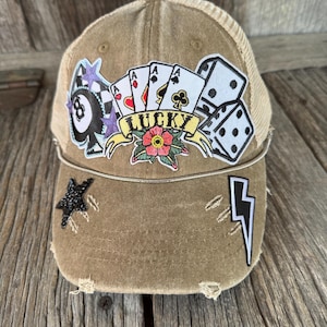 May include: A brown distressed baseball cap with a variety of patches, including playing cards, dice, a star, a lightning bolt, and a tattoo-style patch that says "LUCKY".