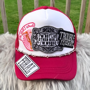 May include: A pink and white trucker hat with multiple patches and a silver chain. Patches include text that reads "The Hell I Won't", "I'm Not Like Other Girls I'm Worse", "Trouble Maker", and "Feral".