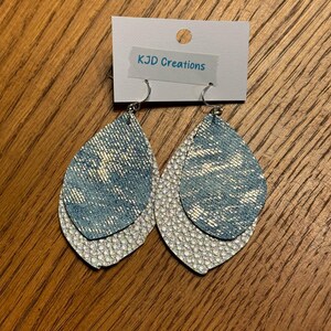 May include: A pair of layered teardrop earrings. The top layer is a light blue denim-like material, and the bottom layer is a textured silver. The earrings hang from silver hooks. The tag above the earrings reads "KJD Creations".