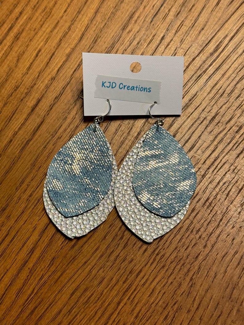 May include: A pair of layered teardrop earrings. The top layer is a light blue denim-like material, and the bottom layer is a textured silver. The earrings hang from silver hooks. The tag above the earrings reads "KJD Creations".