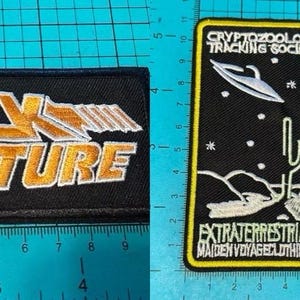 Puede incluir: Dos parches bordados. Uno dice "BACK TO THE FUTURE" en oro y blanco sobre un fondo negro. El otro parche presenta un OVNI, un cactus y el texto "CRYPTOZOOLOGY TRACKING SOCIETY" y "EXTRATERRESTRIALS MAIDEN VOYAGE CLOTHING CO."