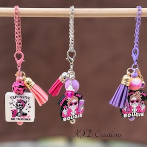 May include: Three decorative charms with pink, silver, and purple chains. Each charm features a skull design with the phrases "EXPENSIVE AND TALKS BACK" and "BROKE AND BOUGIE". The charms include tassels and beads.