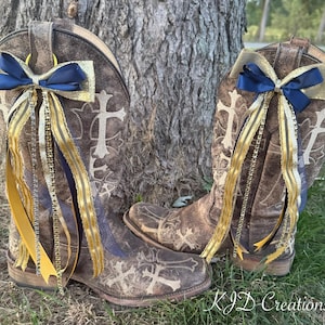 May include: A pair of brown leather cowboy boots with cross designs, adorned with navy blue and gold bows and ribbons. The boots are decorated with gold and navy blue ribbons and are resting on grass.