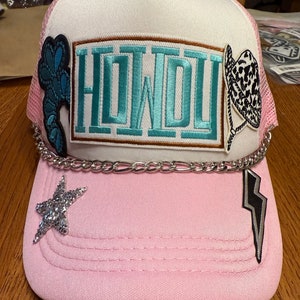 Könnte beinhalten: Ein rosa-weißer Trucker-Hut mit einer silbernen Kette, einem silbernen Stern und einem Blitz-Patch. Der Hut hat einen türkisfarbenen und braunen Patch mit der Aufschrift "HOWDY".