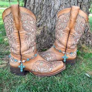 Puede incluir: Un par de botas vaqueras de cuero color canela con intrincados patrones recortados y capas inferiores claras. Cada bota está adornada con una tobillera de cadena plateada con pequeñas cuentas de color turquesa y un colgante en forma de cruz.