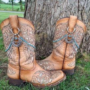 Op de afbeelding: Een paar bruine leren cowboylaarzen met ingewikkelde uitgesneden patronen en gouden onderlagen. Elke laars is versierd met een zilverkleurige hoefijzerbedel, een klein kruisje en een streng turkooiskleurige kralen aan een ring.