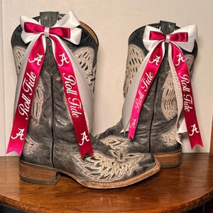Op de afbeelding: Een paar verweerde grijze cowboylaarzen met crèmekleurige borduursels. Elke laars is versierd met een witte strik en een karmozijnrood lint met "Roll Tide" en de letter "A". De laarzen staan op een houten oppervlak. De laarzen zijn ongeveer 30 cm hoog.