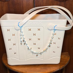 Pode incluir: Uma sacola tote creme com o logotipo Hurley e um charme decorativo de concha e estrela do mar. A sacola tem detalhes recortados e uma alça. A sacola está em uma superfície de madeira.