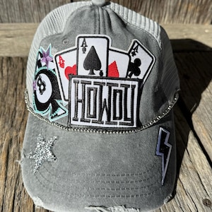 May include: A gray distressed trucker hat with a mesh back. The hat has several patches including playing cards, a 8 ball, a star, and a lightning bolt. The word "HOWDY" is on one of the patches.