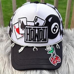 May include: Black and white trucker hat with a mesh back and a curved brim. The hat features patches of playing cards, an 8-ball, dice, and the word "HOWDY". Charms dangle from the brim.