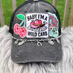 Può includere: Cappellino da camionista grigio slavato con retro in rete nera. Le toppe includono ciliegie, un cappello da cowboy rosa e la scritta "Baby I'm a Wild Card". Una catena di ciondoli argentati con ferri di cavallo e altri ciondoli decora la tesa. Il cappellino ha un aspetto vintage usurato.