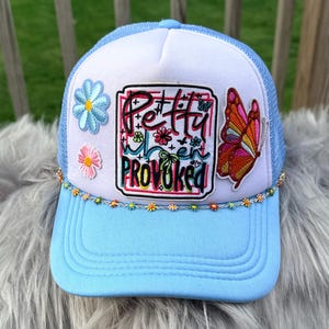 May include: A light blue trucker hat with a white front panel. The hat features embroidered flowers, a butterfly, and the text "Pretty When Provoked". A beaded floral trim accents the brim.