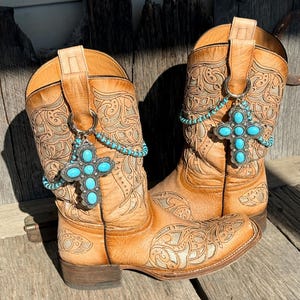 Puede incluir: Un par de botas vaqueras de cuero color canela con intrincados patrones de volutas en relieve. Cada bota está adornada con un colgante de cruz de metal plateado con incrustaciones de piedras turquesas, suspendido de una cadena de cuentas turquesas.