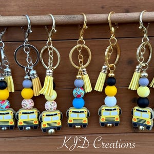 May include: A collection of keychains featuring yellow school bus charms. Each keychain has a beaded design with yellow, white, and patterned beads, a yellow tassel, and a metal clasp. The text "KJD Creations" is visible.