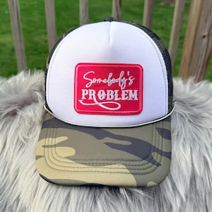 Op de afbeelding: Truckerpet met een wit voorpaneel en een camouflageklep. De pet heeft een rode rechthoekige patch met de woorden "Somebody's PROBLEM" in witte letters.