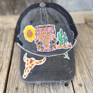 May include: Distressed black trucker hat with a black mesh back. The hat is decorated with patches including a sunflower, a steer skull, a cactus, and the word "COWGIRL". A silver chain with a turquoise charm hangs from the brim.