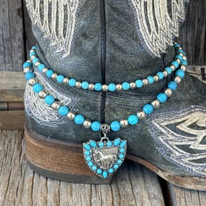 May include: A turquoise and silver beaded necklace with a shield pendant featuring a horse, resting on a black cowboy boot with decorative stitching. The necklace is composed of small, round turquoise and silver beads.