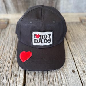 May include: Black baseball cap with a red heart patch on the brim and a rectangular patch that reads "I ❤️ HOT DADS" in white letters with a red heart. The hat is made of a durable fabric.