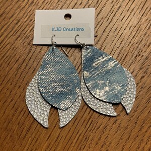 May include: A pair of layered teardrop earrings. The top layer is denim-look blue with silver accents, and the bottom layer is white with a textured, scale-like pattern. The earrings hang from silver hooks. The tag reads "KJD Creations".