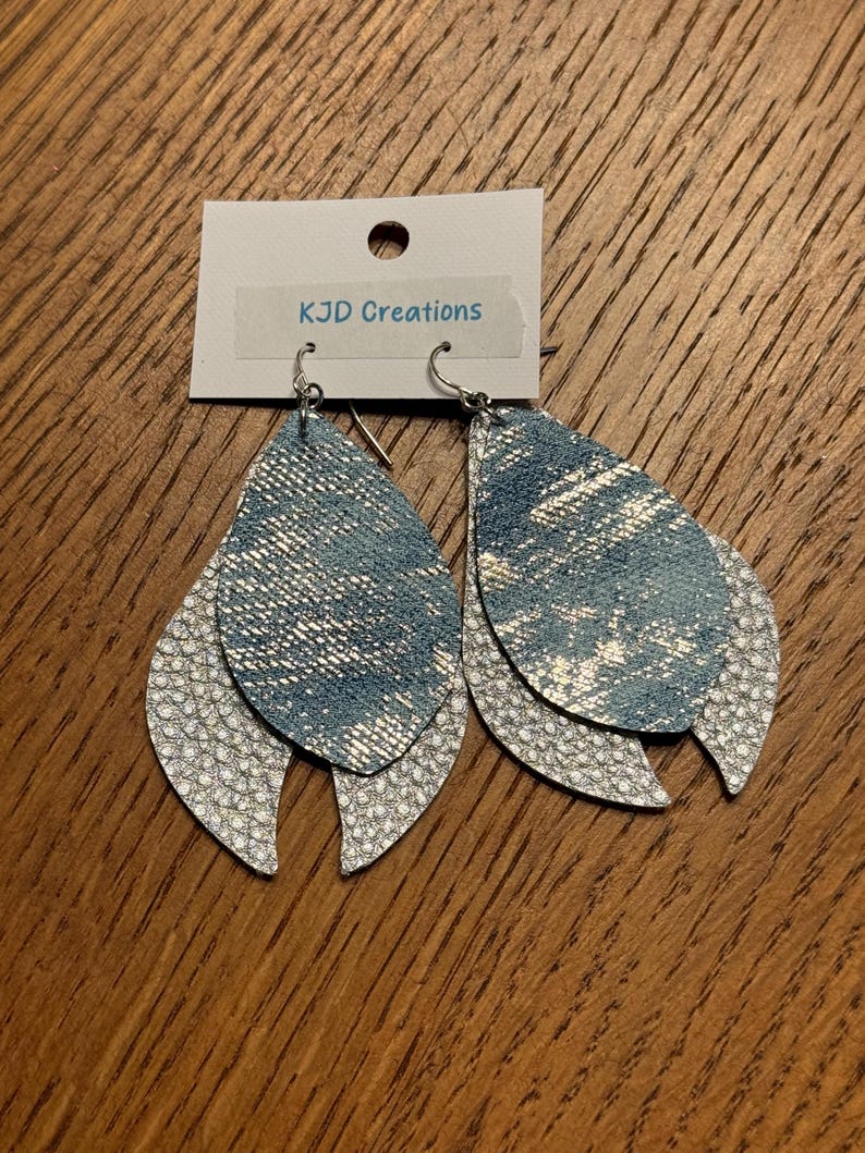 May include: A pair of layered teardrop earrings. The top layer is denim-look blue with silver accents, and the bottom layer is white with a textured, scale-like pattern. The earrings hang from silver hooks. The tag reads "KJD Creations".