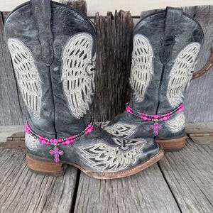 Puede incluir: Un par de botas de vaquero de cuero negro desgastado con diseños de alas bordadas y una pulsera de tobillo con un dije de cruz rosa. Las botas tienen punta cuadrada y tacón apilado.