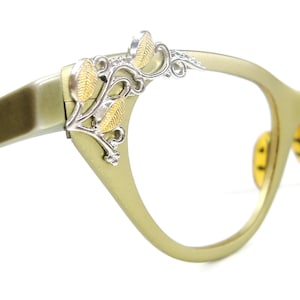 May include: Cat-eye glasses with a matte olive green frame and a silver-toned metal embellishment on the temple corner. The decorative piece features a vine-like design with textured gold-colored leaves and small silver swirls.