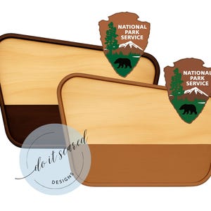 May include: Two wooden plaques with a light wood grain top and a dark brown and tan bottom. Each plaque has a National Park Service logo with a bear, mountains, and trees. The plaques are in a diagonal orientation.