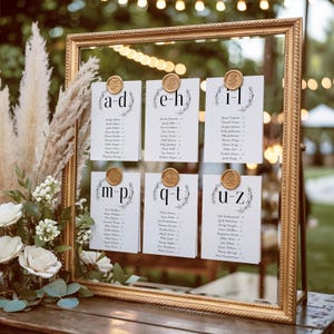 May include: A gold framed seating chart with six white cards, each held by a gold wax seal. The cards are labeled with letters and feature guest names. The frame is on a wooden table with floral arrangements.