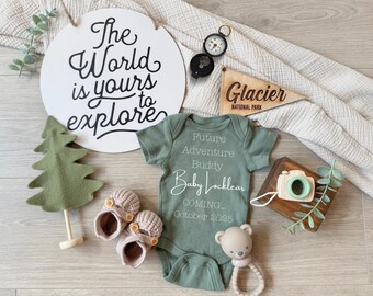 May include: A green baby onesie with the text "Future Adventure Buddy Baby Locklear Coming... October 2025". The onesie is surrounded by a wooden sign that says "Glacier National Park", a small wooden camera, a green felt tree, and a wooden sign with the text "The World is yours to explore".