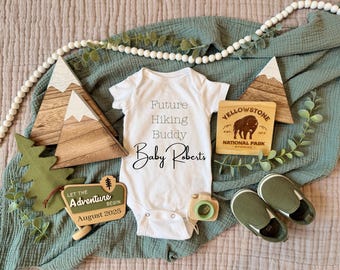 May include: A white onesie with the text "Future Hiking Buddy Baby Roberts" on it. The onesie is surrounded by wooden mountain decorations, a green felt tree, a wooden sign that says "Let the Adventure Begin August 2025", and a wooden sign with the text "Yellowstone National Park".