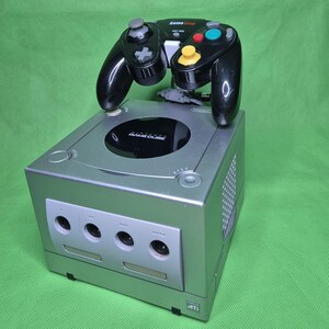 Könnte beinhalten: Eine silberne Nintendo GameCube Spielkonsole mit einem schwarzen GameStop-Controller oben drauf. Die Konsole verfügt über einen runden Deckel mit dem Nintendo GameCube-Logo, vier Controller-Anschlüsse an der Vorderseite und ein ATI-Logo.