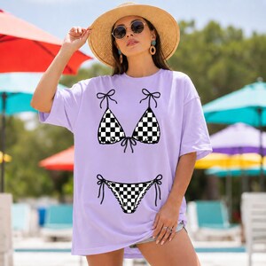 以下が含まれることがあります： 黒と白のチェック柄ビキニのデザインが施された、薄紫色のTシャツ。 ビキニのトップスとボトムは、リボンのディテールで描かれています。 このシャツはカジュアルなオーバーサイズスタイルです。