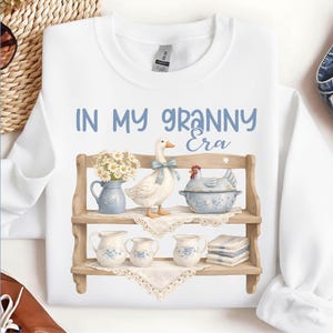 May include: White sweatshirt with the text "IN MY GRANNY Era" in blue. The design features a wooden shelf with a goose, a chicken, and floral-decorated pitchers and bowls. A charming, vintage-inspired design.