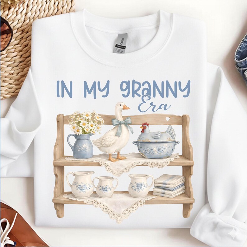 May include: White sweatshirt with the text "IN MY GRANNY Era" in blue. The design features a wooden shelf with a goose, a chicken, and floral-decorated pitchers and bowls. A charming, vintage-inspired design.
