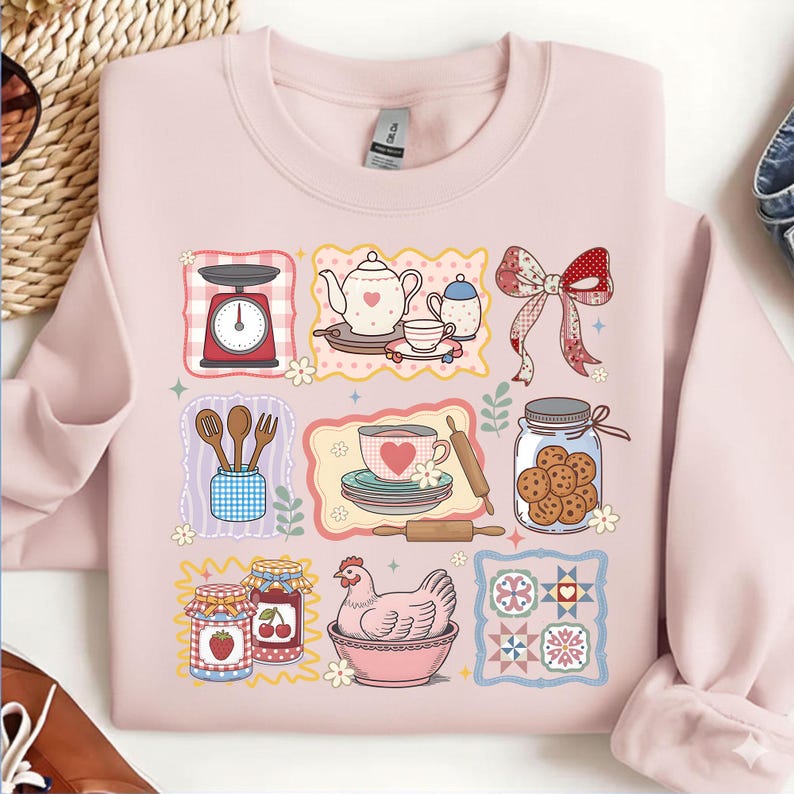May include: A light pink sweatshirt featuring a collection of illustrated kitchen and baking elements. The design includes a scale, teapot, teacup, cookies, rolling pin, and a chicken in a bowl. The graphics are in a cartoon style.