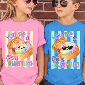 Puede incluir: Dos niños vistiendo camisetas rosas y azules con un gráfico de dumpling. La camiseta rosa tiene un dumpling con un lazo, y la camiseta azul tiene un dumpling con gafas de sol. Ambas camisetas dicen "You're a Glitter Dumpling."