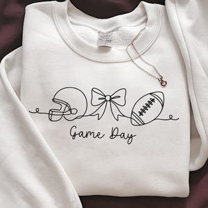 May include: White sweatshirt with a black line drawing of a football helmet, bow, and football. The words "Game Day" are written below the drawing in a script font. A delicate necklace with a small pendant is draped on the sweatshirt.