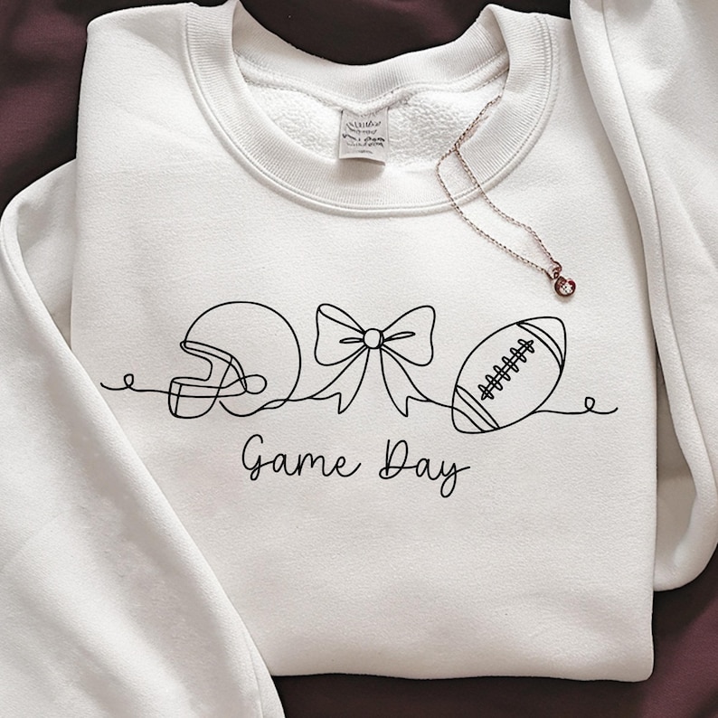 May include: White sweatshirt with a black line drawing of a football helmet, bow, and football. The words "Game Day" are written below the drawing in a script font. A delicate necklace with a small pendant is draped on the sweatshirt.