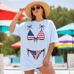 以下が含まれることがあります： アメリカ国旗風のビキニデザインが施された水色のTシャツ。ビキニのトップスとボトムは、赤と白のストライプと、白星の青で描かれています。麦わら帽子とサングラスをかけた人が着ています。