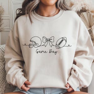 May include: A cream-colored sweatshirt featuring a black line drawing of a football helmet, a bow, and a football. The words "Game Day" are written in a script font below the drawings. The sweatshirt is a casual style.