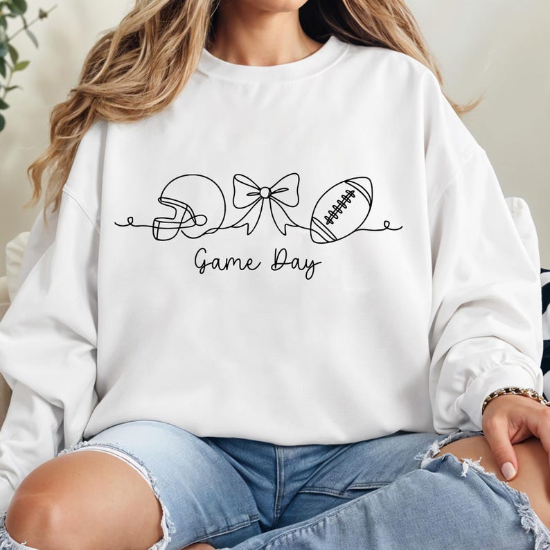 May include: White sweatshirt with a black line drawing of a football helmet, bow, and football. The words "Game Day" are written below the drawing in a script font. The sweatshirt is a casual style.