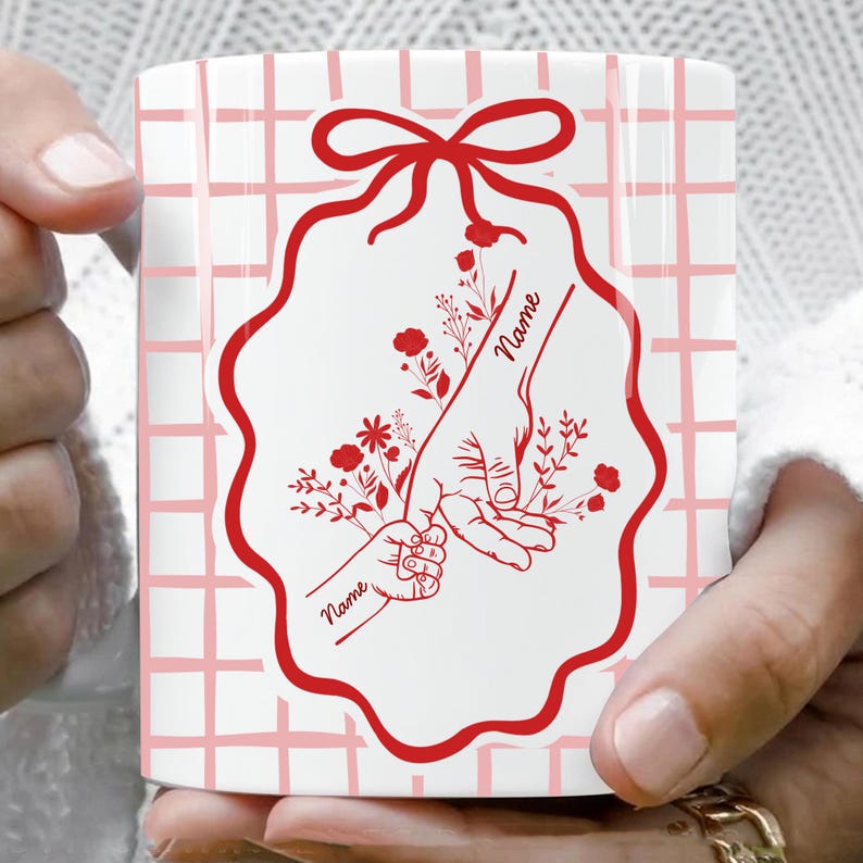 May include: White ceramic mug with a red floral design featuring a parent's hand holding a child's hand, with the word "Name" in script. The mug has a pink grid pattern background and a red bow at the top.