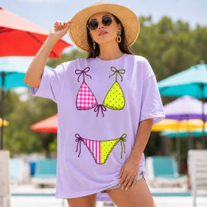 以下が含まれることがあります： 遊び心のあるビキニのデザインが特徴の、薄紫色のTシャツ。ビキニトップはピンクのギンガムチェック柄と黄色の水玉模様で描かれています。ビキニボトムはピンクと白のストライプ柄と黄色の水玉模様です。