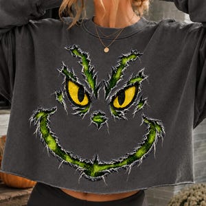Könnte beinhalten: Ein anthrazitfarbenes, langärmeliges bauchfreies Sweatshirt mit einem abgenutzten, ausgefransten Grafikdruck eines grünen Gesichts mit leuchtend gelben Augen und einem breiten, schelmischen Grinsen. Das Design wirkt strukturiert und zerrissen.