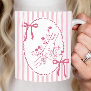May include: White ceramic mug with pink vertical stripes and a pink oval design. The oval features a parent's hand holding a child's hand, with floral accents and the word "Name". Pink bows adorn the top and bottom.
