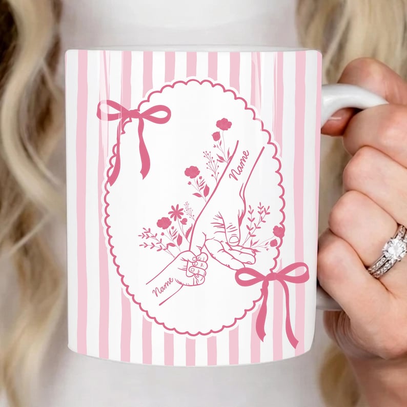 May include: White ceramic mug with pink vertical stripes and a pink oval design. The oval features a parent's hand holding a child's hand, with floral accents and the word "Name". Pink bows adorn the top and bottom.